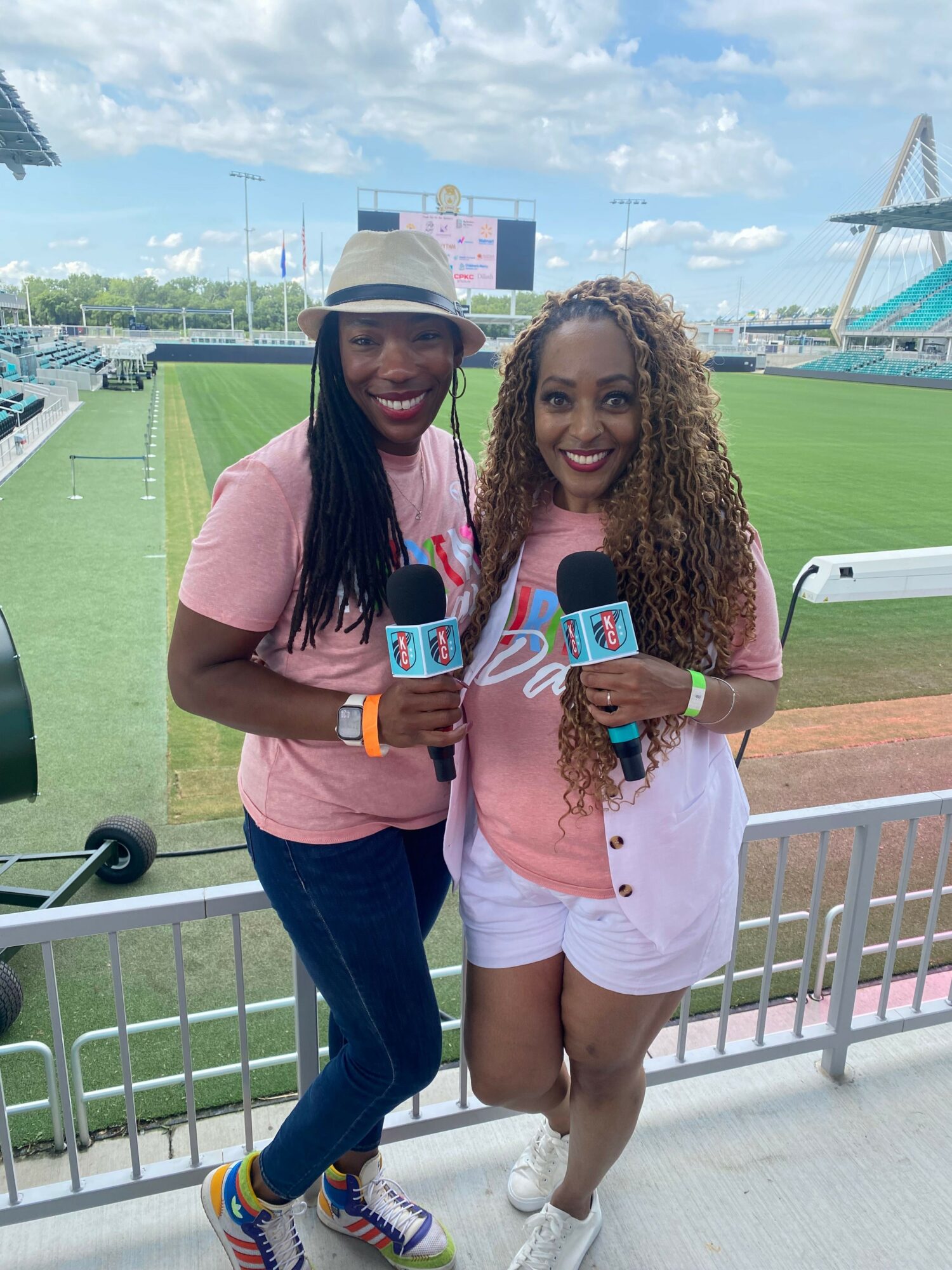 Two women standing outdoors near a railing, holding microphones, with a sports field and buildings in the background.