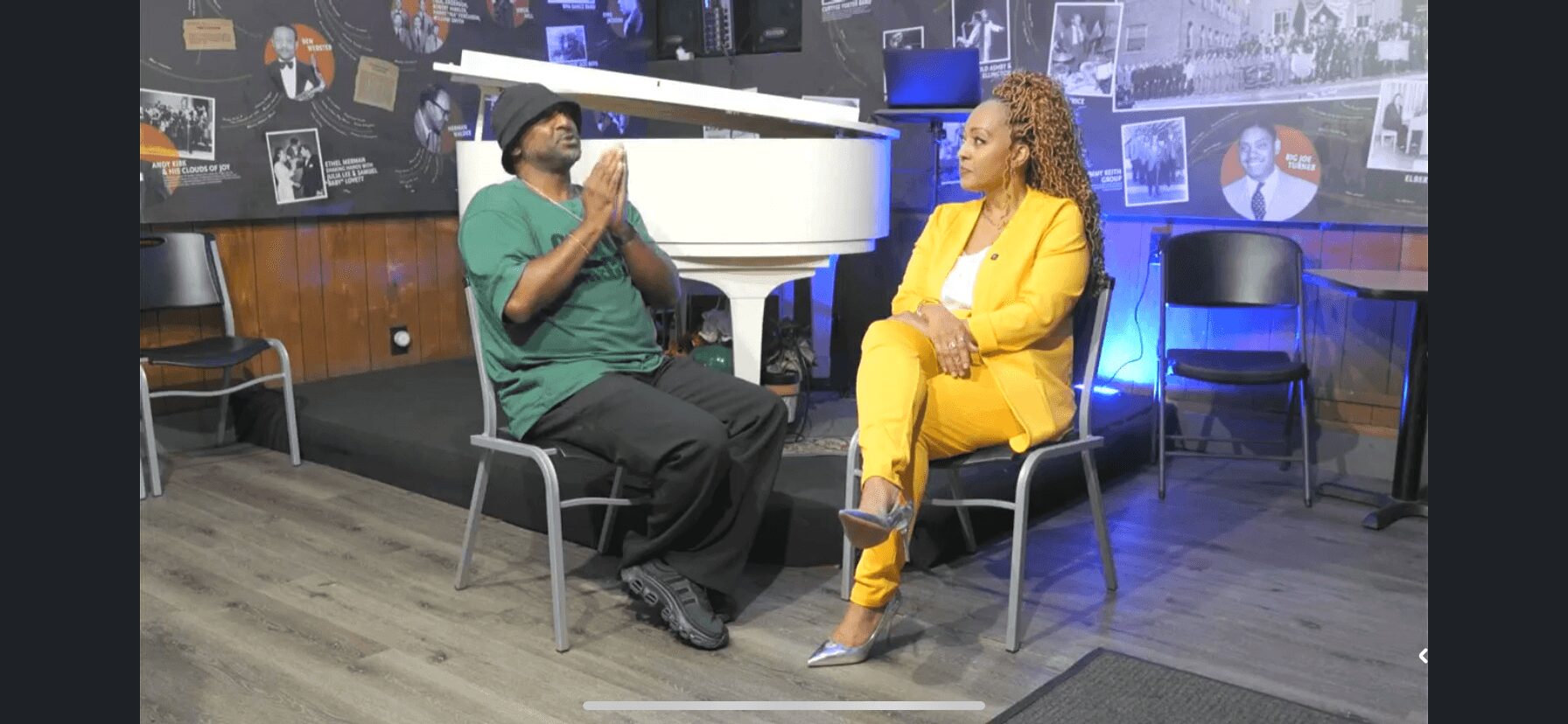 Two people seated and talking in a studio with photos on the wall, one in green and the other in yellow, on chairs.
