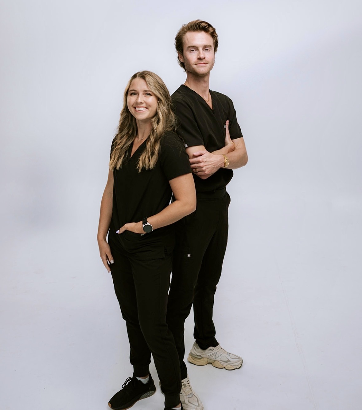 Two people standing side by side, smiling, with a plain light background.