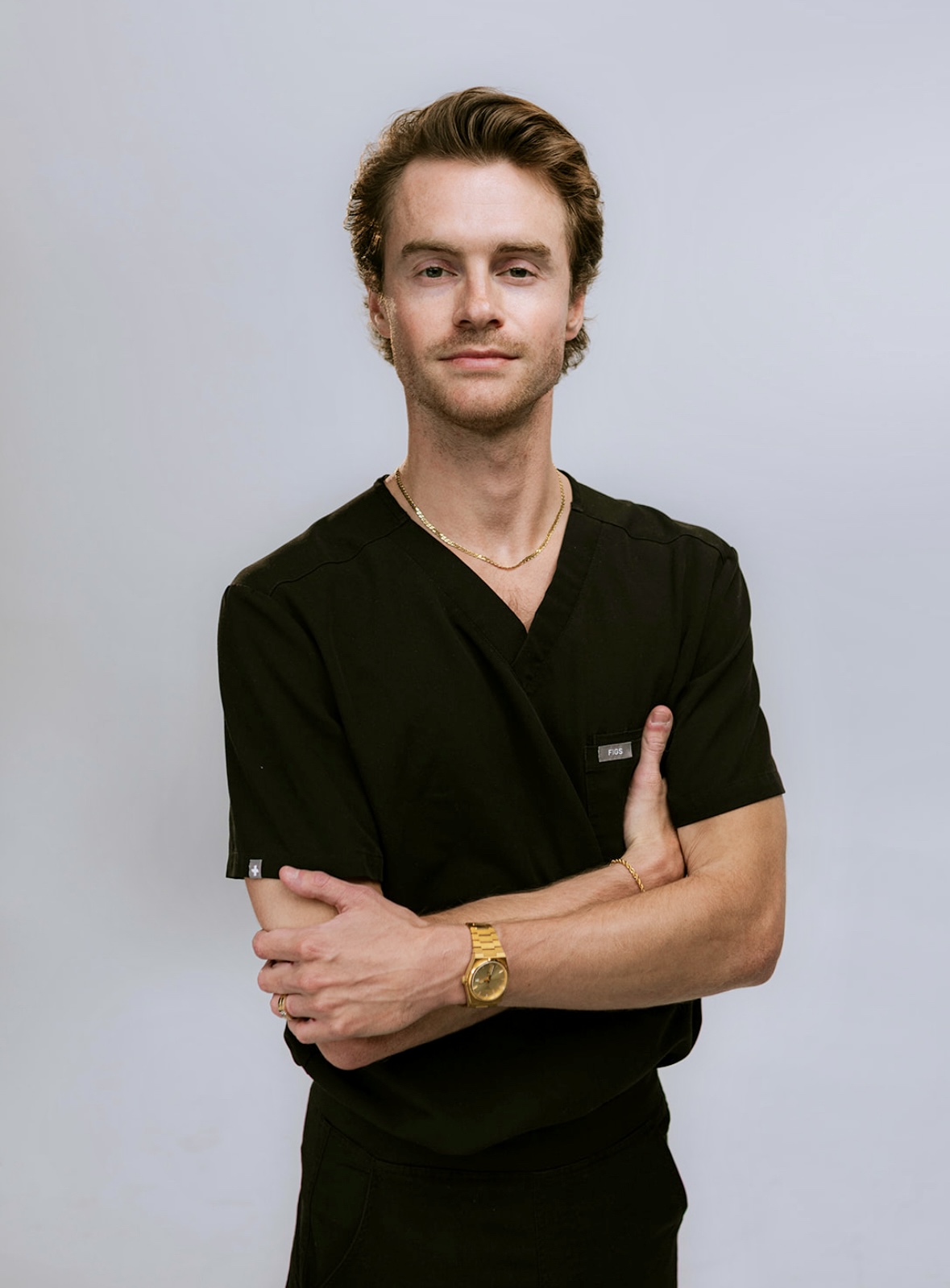 Young man with light brown hair, wearing black scrubs, gold watch, and necklace, standing with arms crossed against a light background.