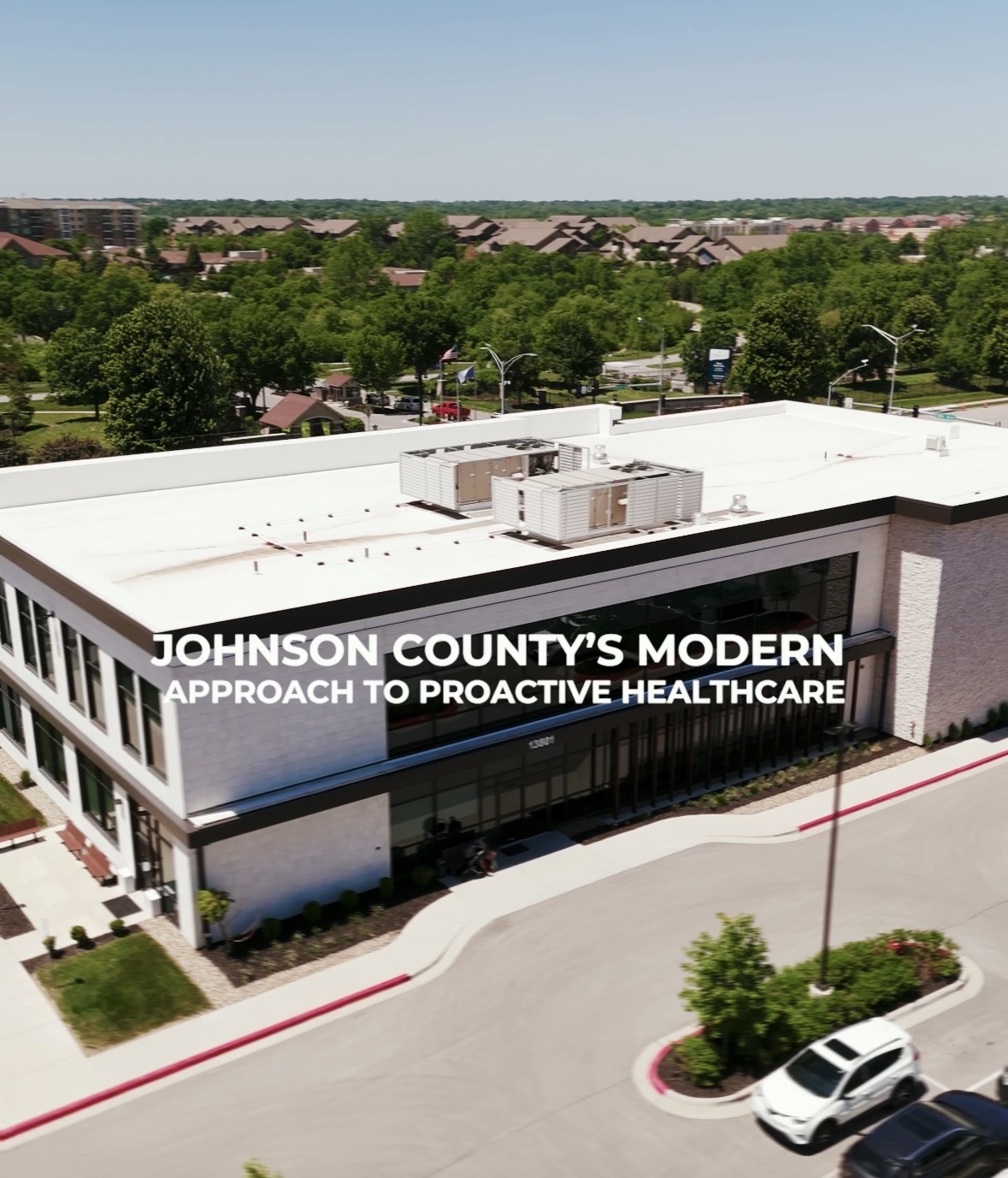 Modern healthcare building with white roof, surrounded by trees and a parking lot, under a clear sky.