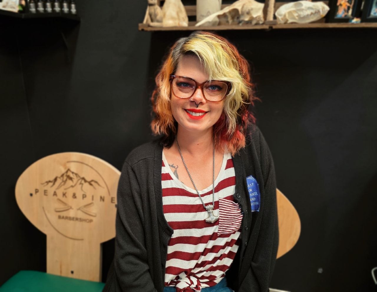Smiling woman with colorful hair and glasses, wearing a striped shirt and black cardigan, sitting indoors.