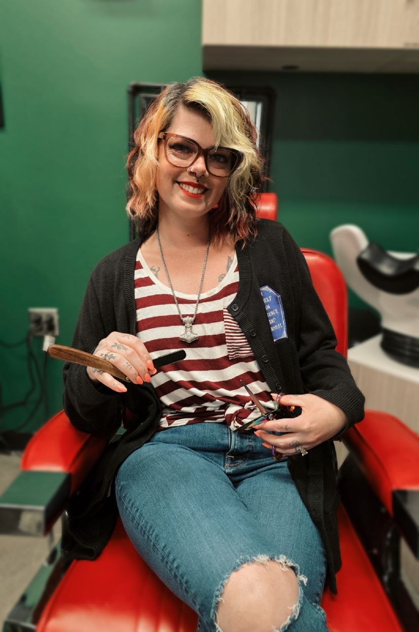 Person with curly blonde hair and glasses smiling, sitting in a chair, holding a phone, wearing a striped shirt and jeans.