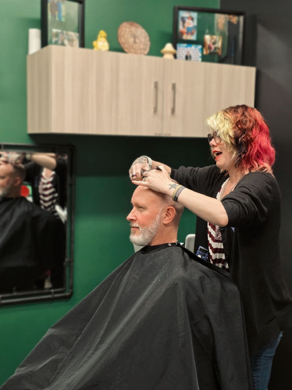 A woman with colorful hair styles a man's hair in a salon, with a mirror and cabinet in the background.