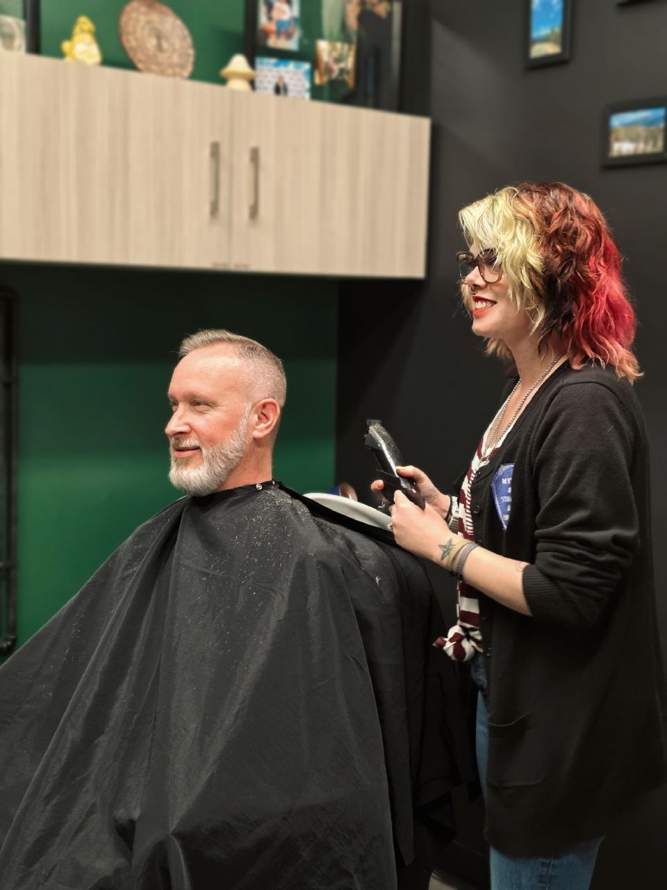 A person with a beard and short hair is sitting in a barber chair, covered with a black cape, smiling. A woman with glasses and colorful hair is standing nearby, holding a phone and smiling.