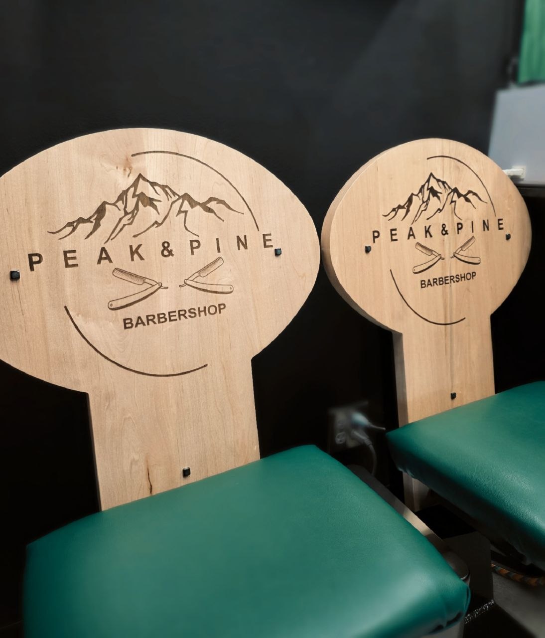 Two wooden chairs with round backs featuring mountain and pine tree illustrations, and the text 'PEAK & PINE BARBERSHOP'.