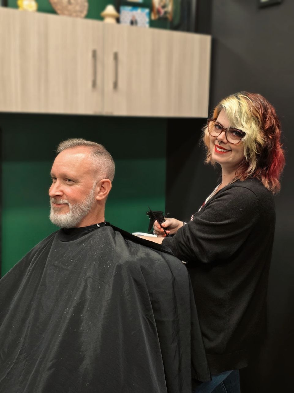 A woman with glasses and colorful hair stands behind a man with a beard in a barber chair, smiling, holding a phone.