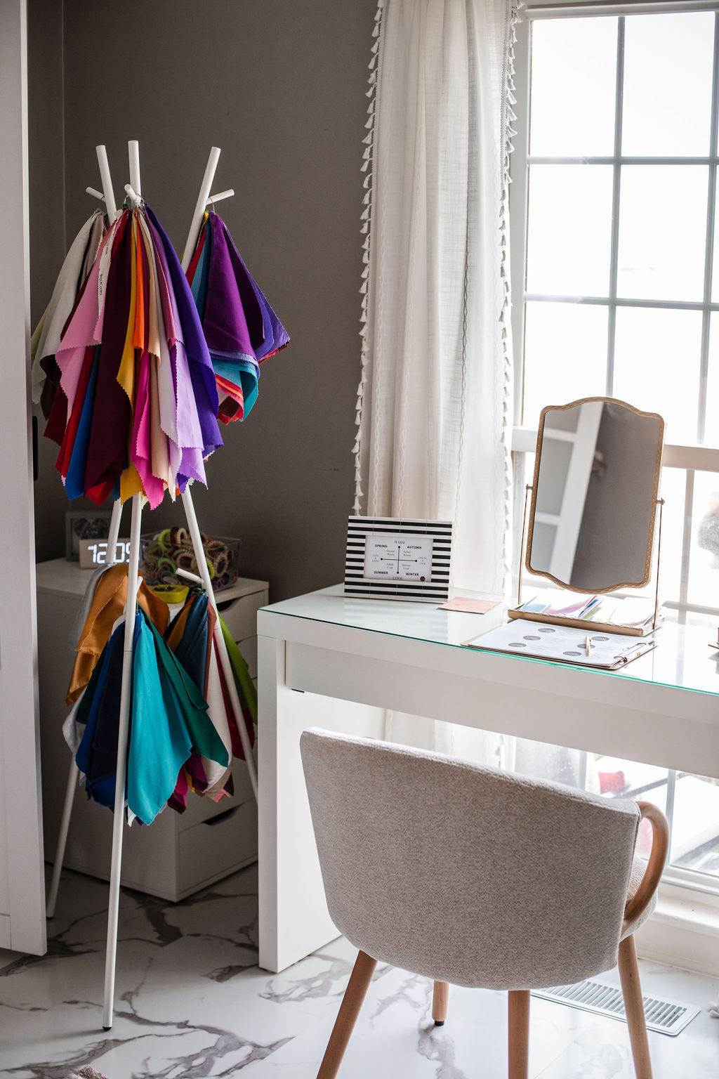 Colorful fabric swatches hanging on a stand next to a white desk with a mirror and a window with curtains.