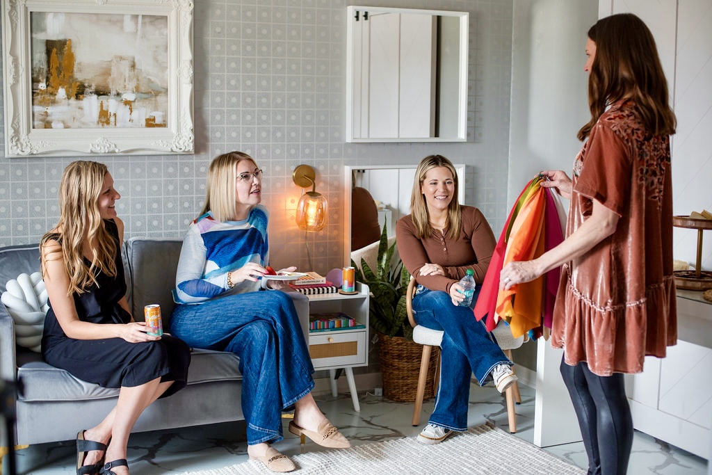 Four women sitting on a sofa and chair in a living room, engaging in conversation with a standing woman holding colorful clothes.