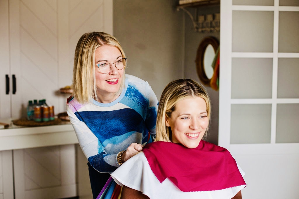 Two women smiling and laughing in a room, one with blonde hair and glasses, the other with blonde hair and a pink cape.