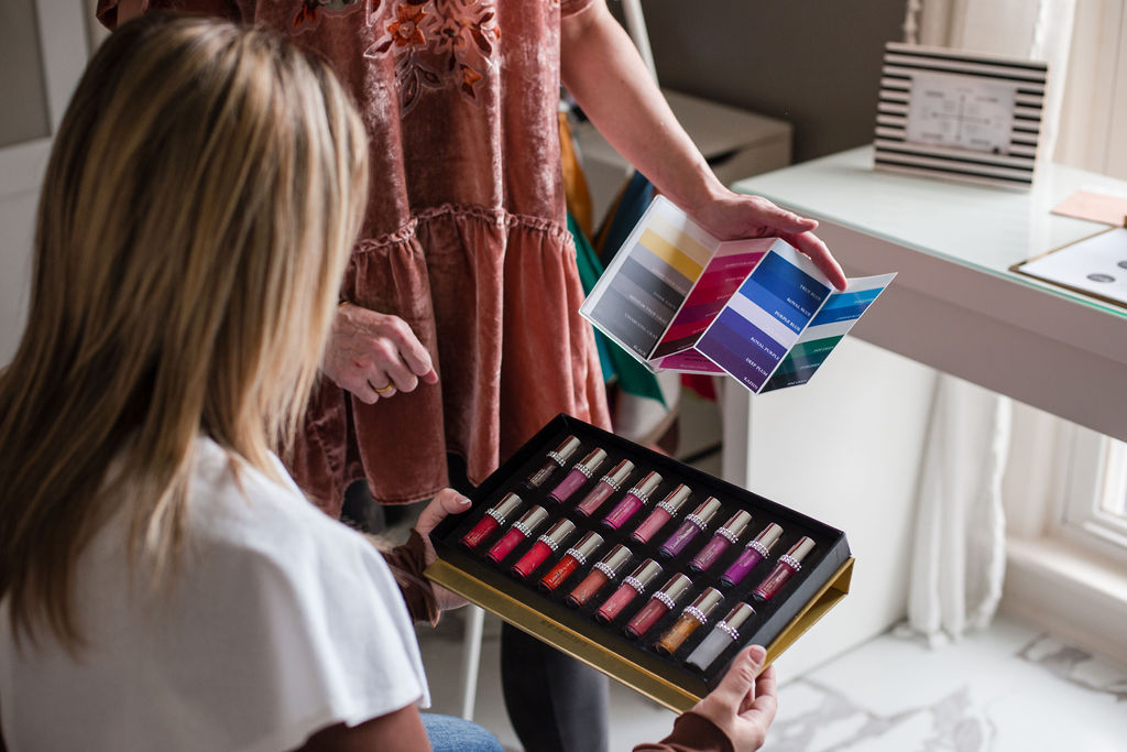 Two women examine a box of colorful nail polish bottles in a bright room with a window and desk.