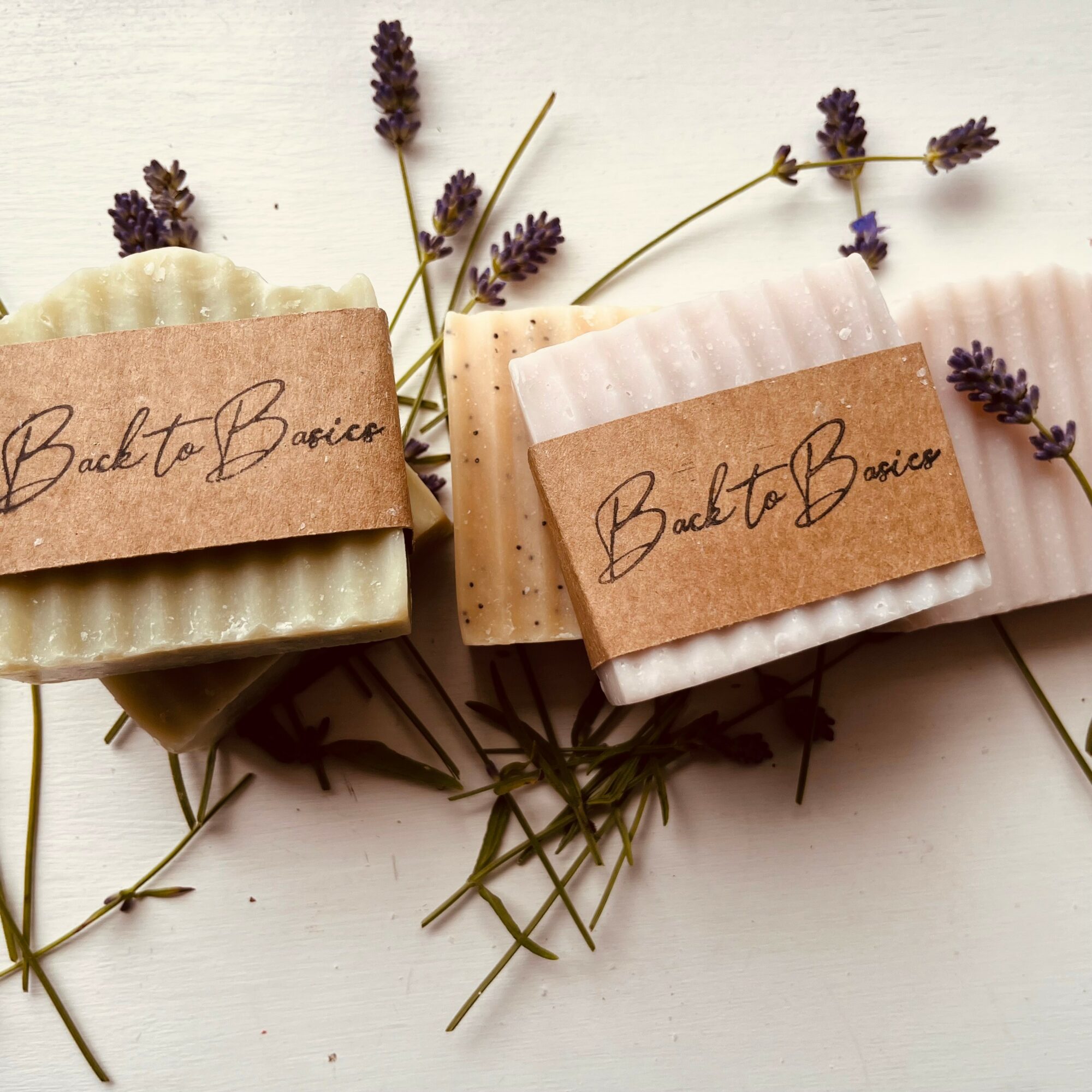 Three handmade soap bars with brown paper labels reading 'Back to Basics,' surrounded by lavender sprigs on a white surface.