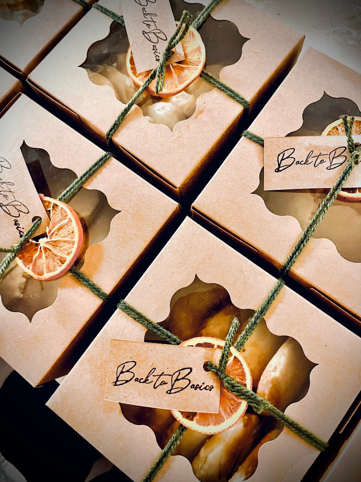 Four gift boxes with windowed lids, tied with green string, decorated with dried orange slices and handwritten tags.
