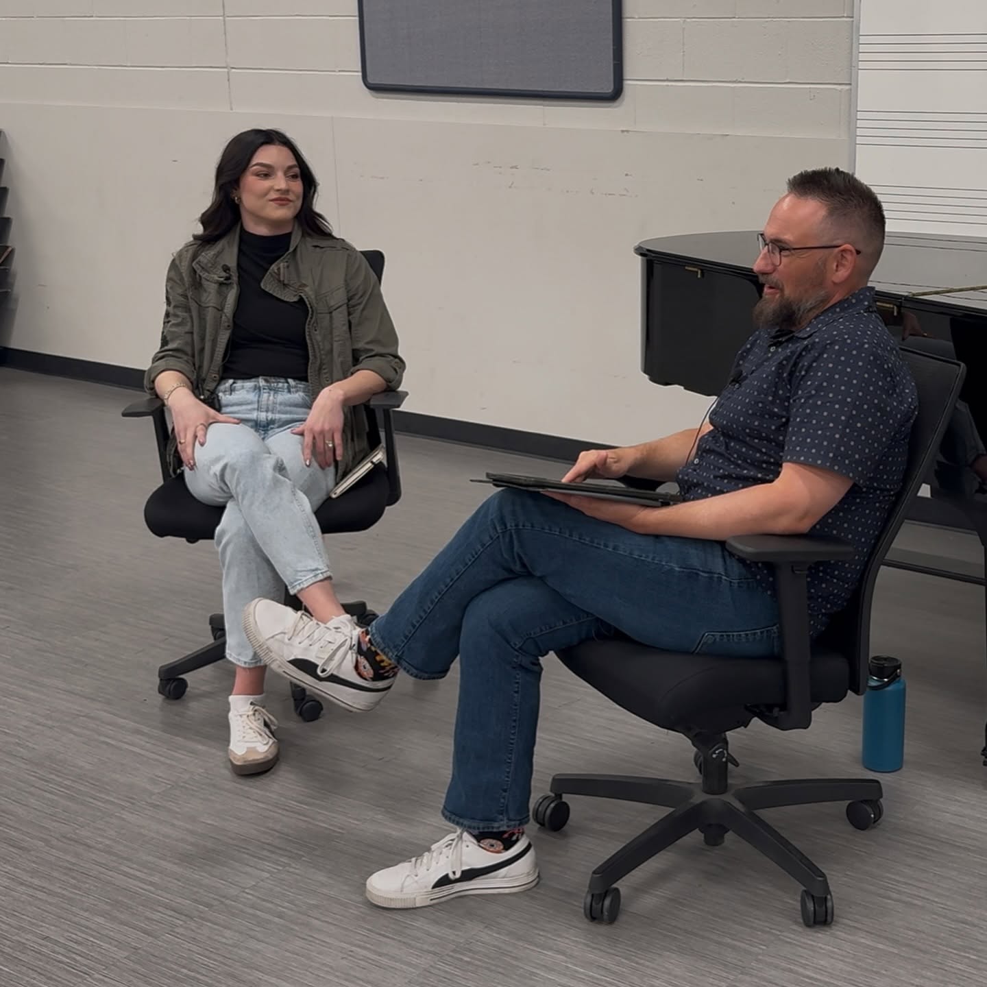 Two people sitting and talking in a room with a white wall and a blackboard, one woman and one man, both smiling.