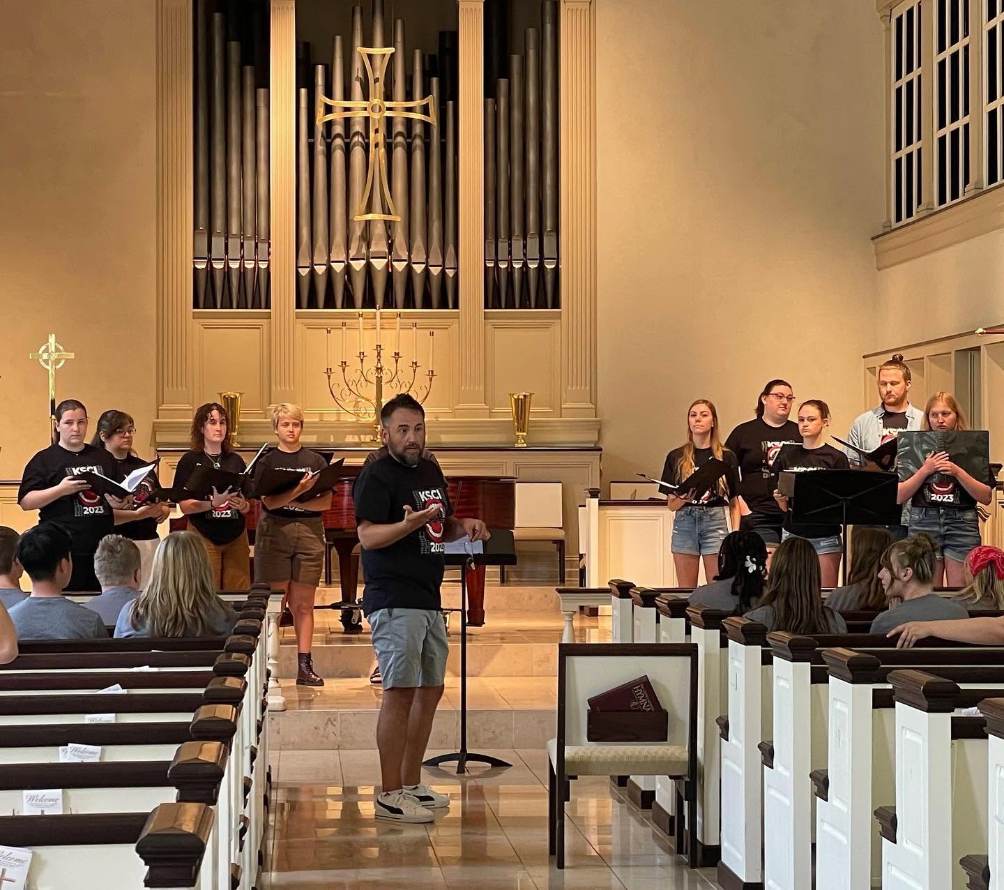 Group of people singing in a church with an organ and candles, audience seated in pews, man in front holding a microphone.