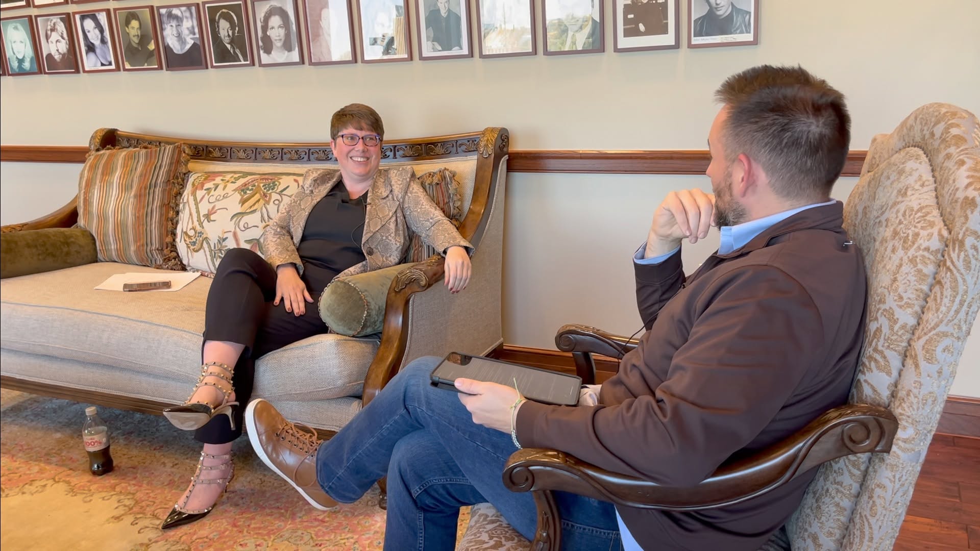 Two people sitting and talking in a room with framed photos on the wall, one on a sofa and the other in a chair.