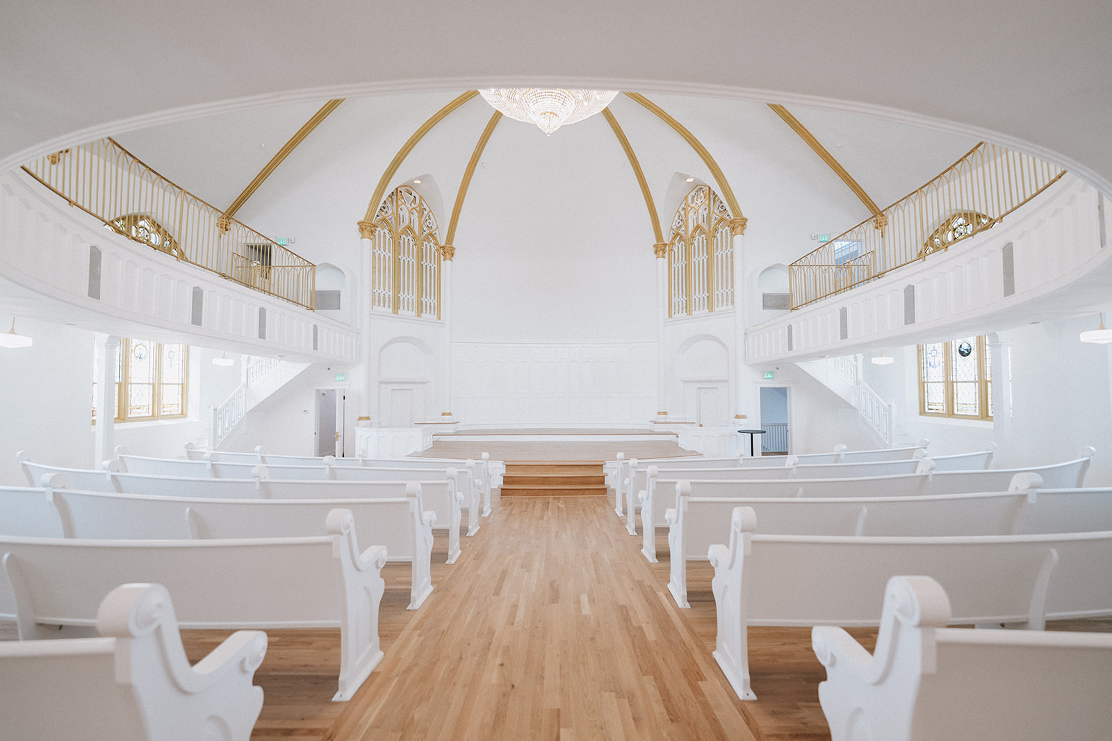 Interior of a bright, white church with wooden floor, white pews, and a high arched ceiling with gold accents.