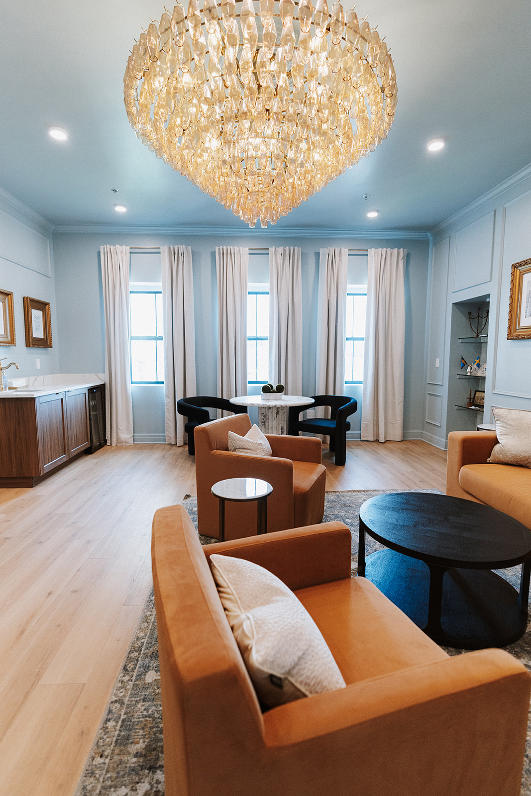 Elegant living room with a large chandelier, light blue walls, tall windows with white curtains, and various seating options.