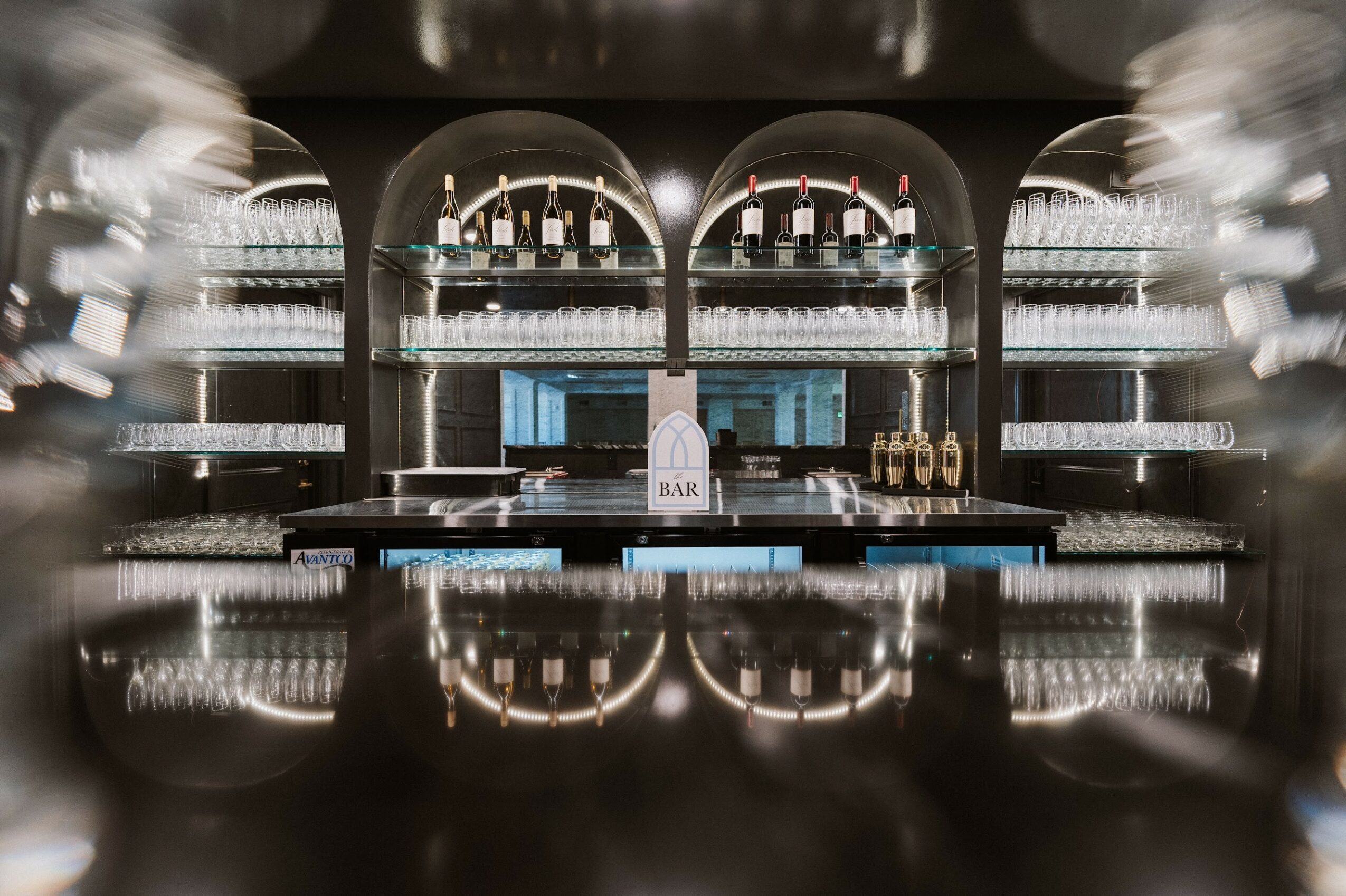 Bar with bottles on shelves, illuminated, with a reflective surface in front, symmetrical arrangement, modern design.