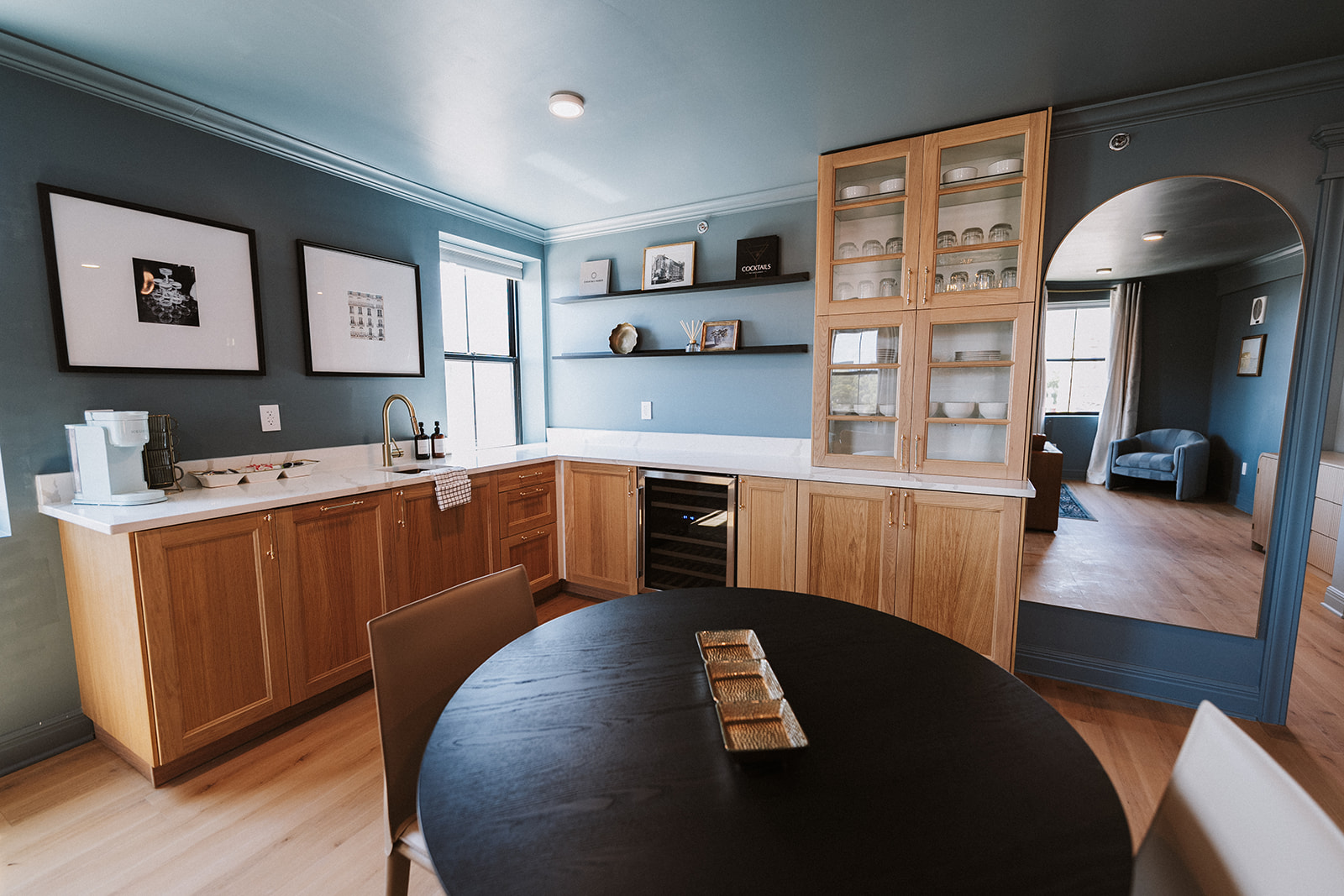 Kitchen with blue walls, wooden cabinets, black countertop, and a round black dining table with a decorative item, adjacent to a living room.