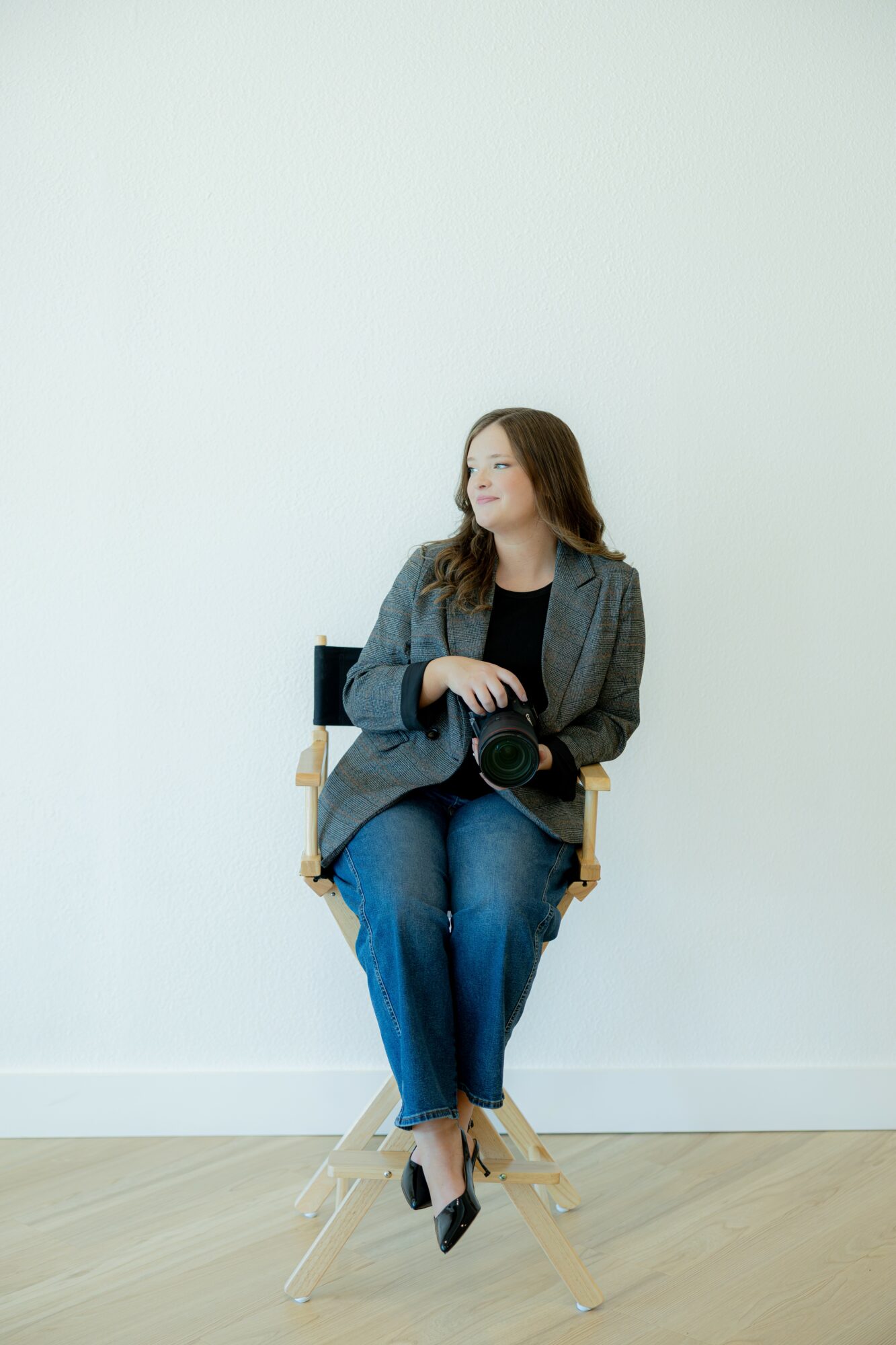 Woman sitting on a chair holding a camera, wearing a blazer and jeans, against a plain white wall.