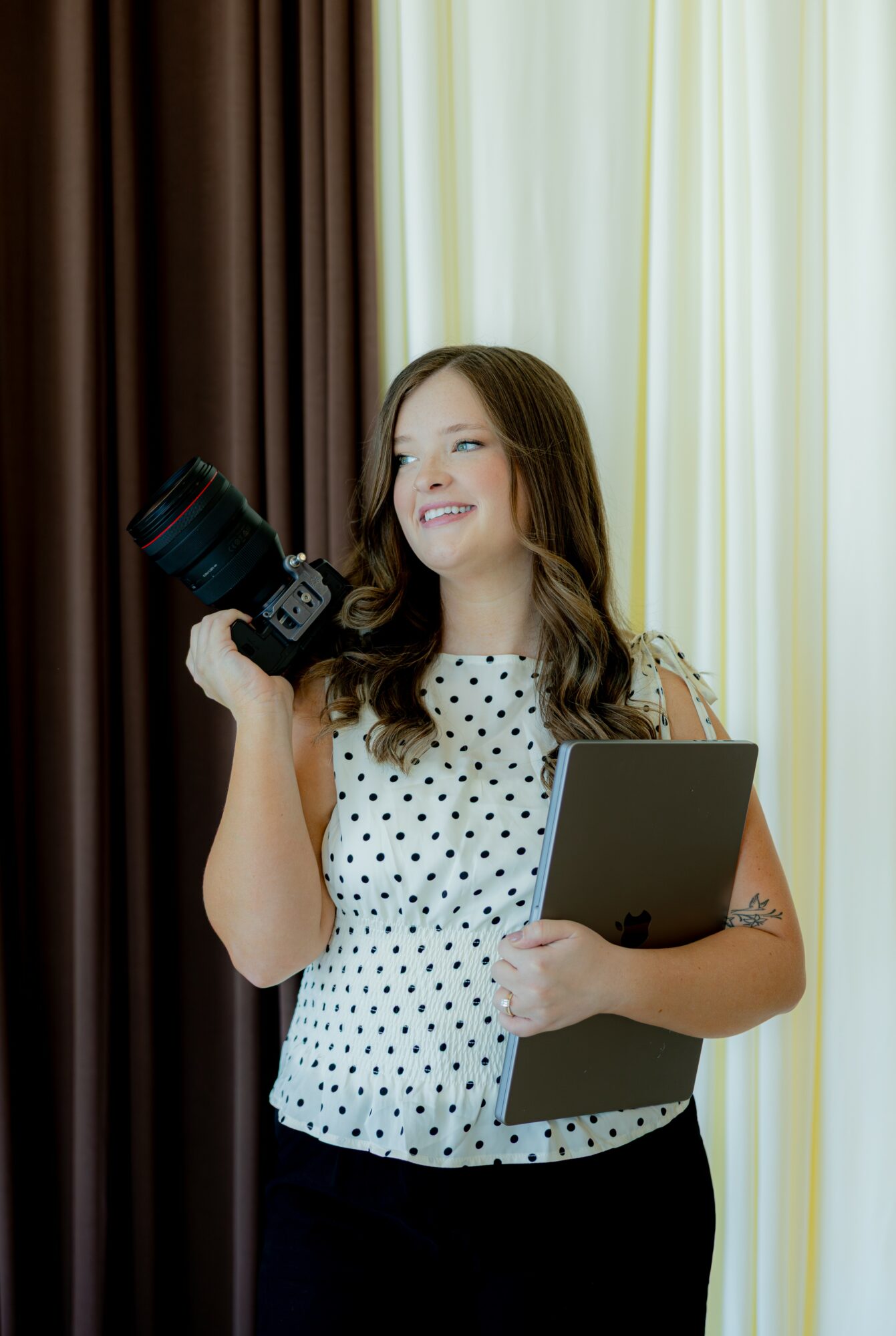Young woman holding a camera and a folder, smiling, standing in front of curtains.