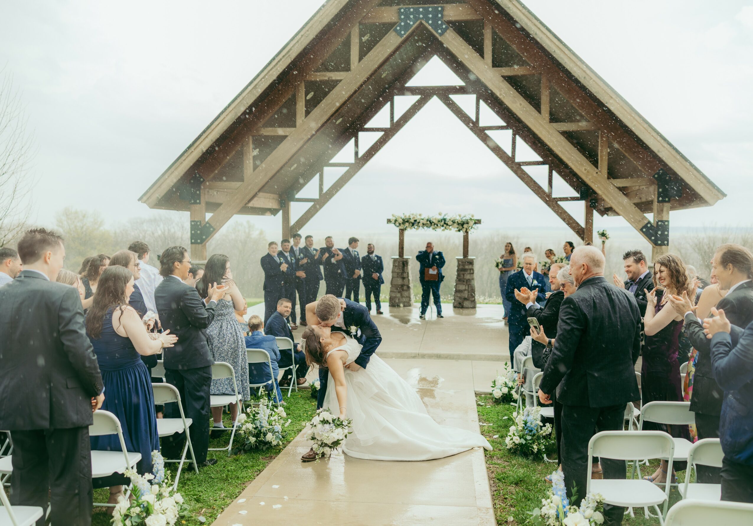 Wedding ceremony outdoors under a wooden A-frame structure with guests standing and sitting, bride walking down aisle, overcast sky.