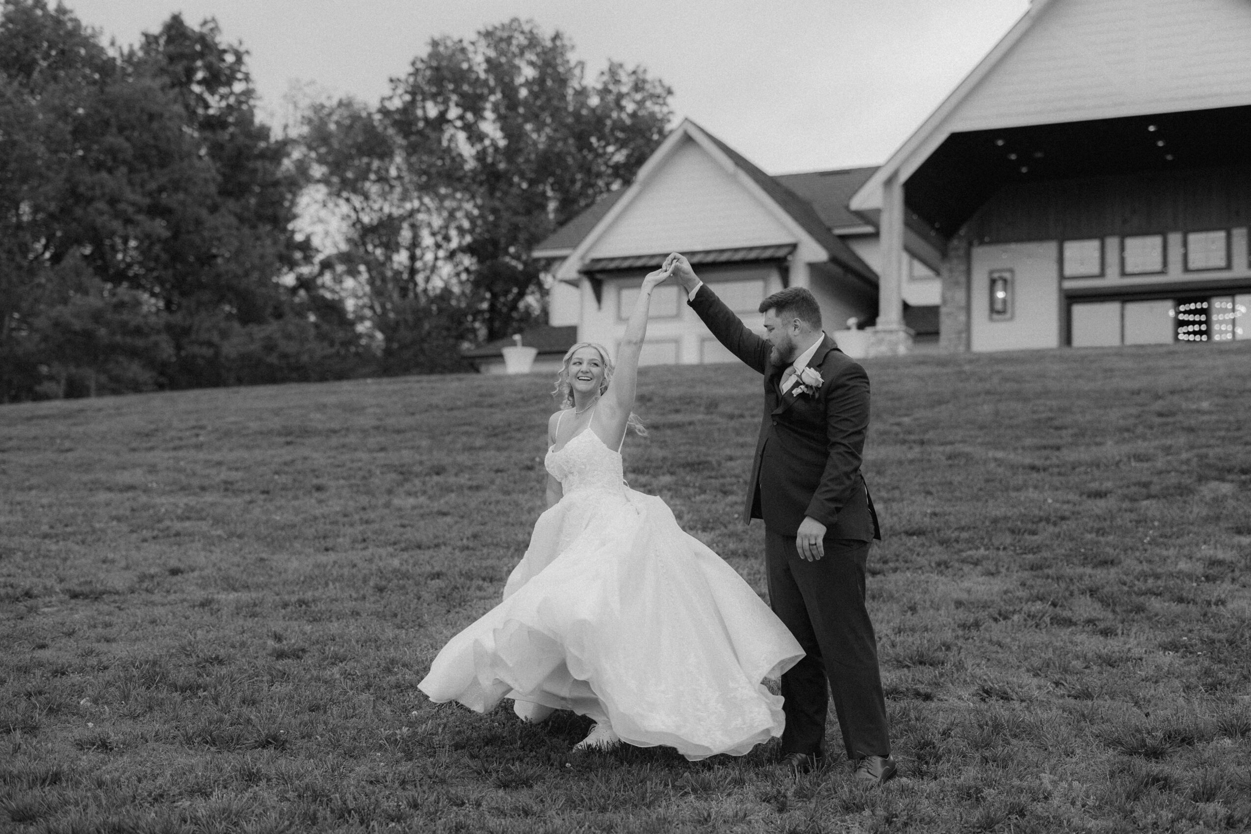 A bride and groom dance outdoors on a grassy hill, with a building and trees in the background.