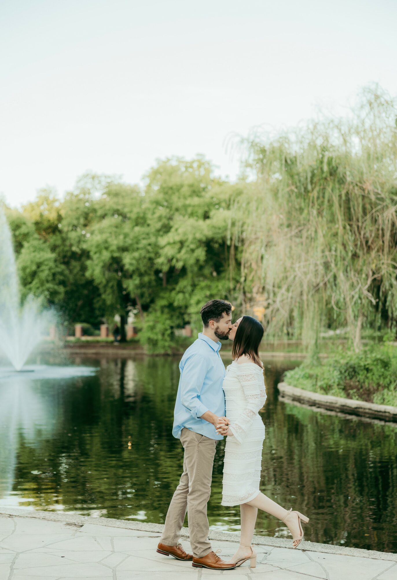 A couple stands by a pond with trees and a fountain in the background, facing each other closely outdoors.
