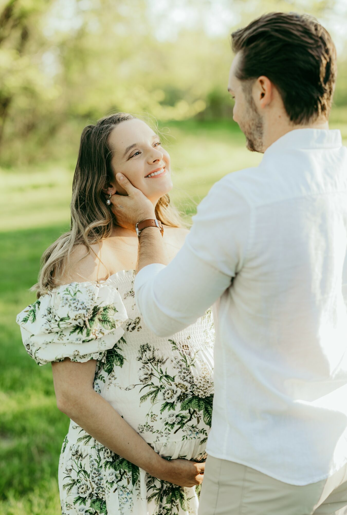 A couple outdoors, the woman smiling and looking at the man, holding her pregnant belly, with trees in the background.