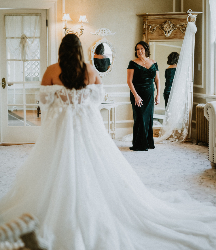 Two women in a room, one in a wedding dress, the other in a dark gown, facing each other, mirror, and vintage decor.