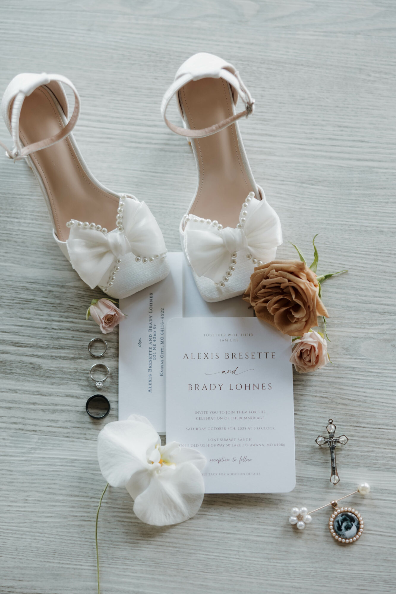 Pair of white high-heeled shoes with bows, a wedding invitation, a flower, jewelry, and a rosary on a light wooden surface.