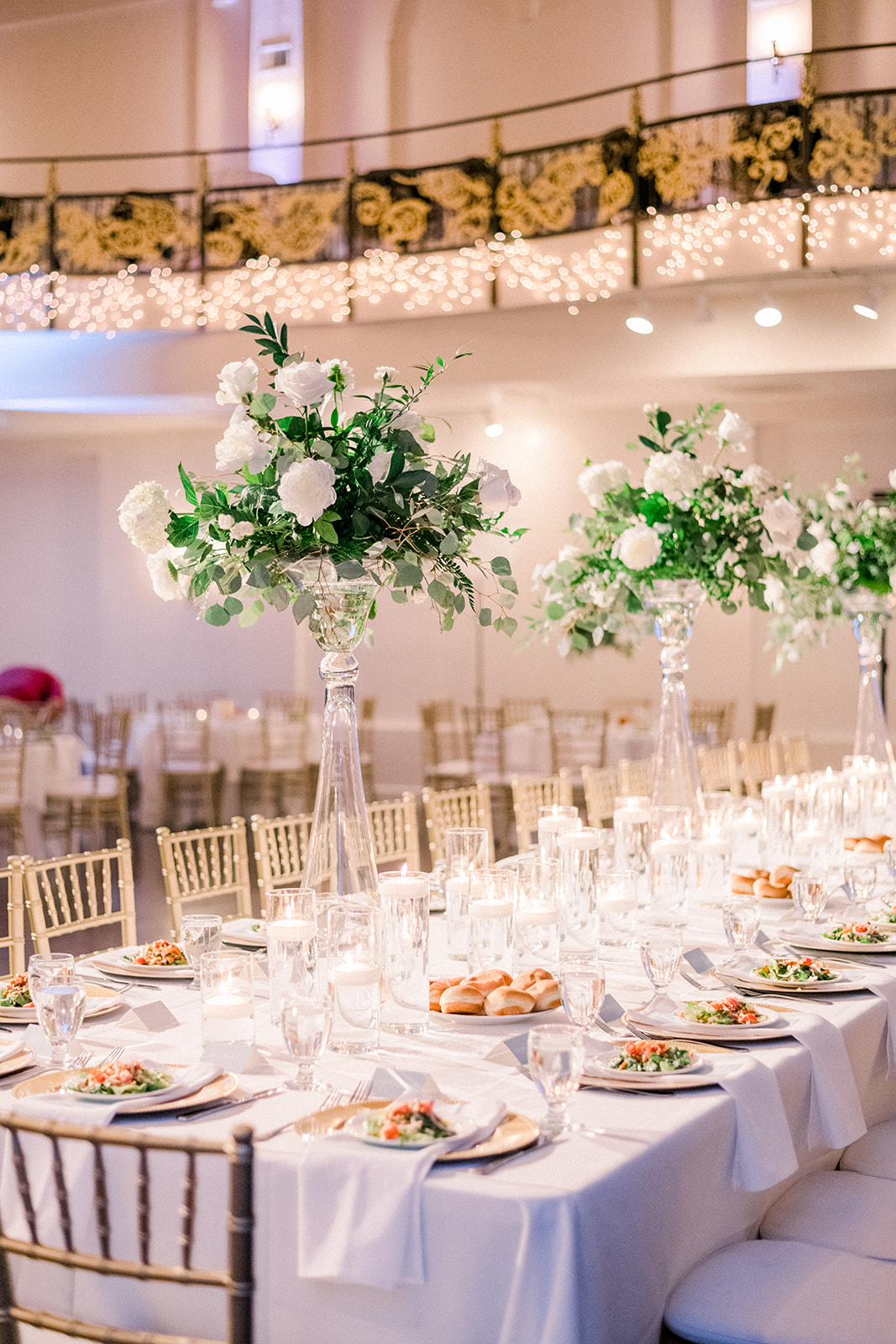Elegant banquet table with tall floral centerpieces, set with plates, glasses, and silverware in a decorated event hall.
