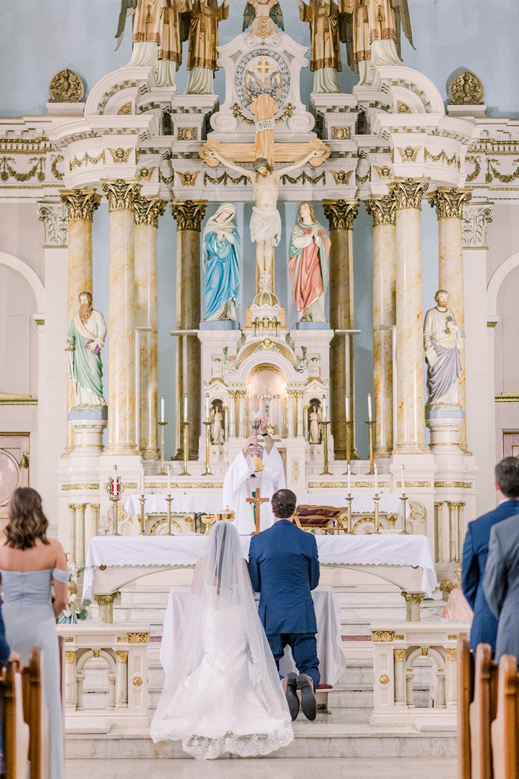 Wedding ceremony at church altar with bride, groom, priest, and guests, ornate gold and white decor, religious statues, and altar.
