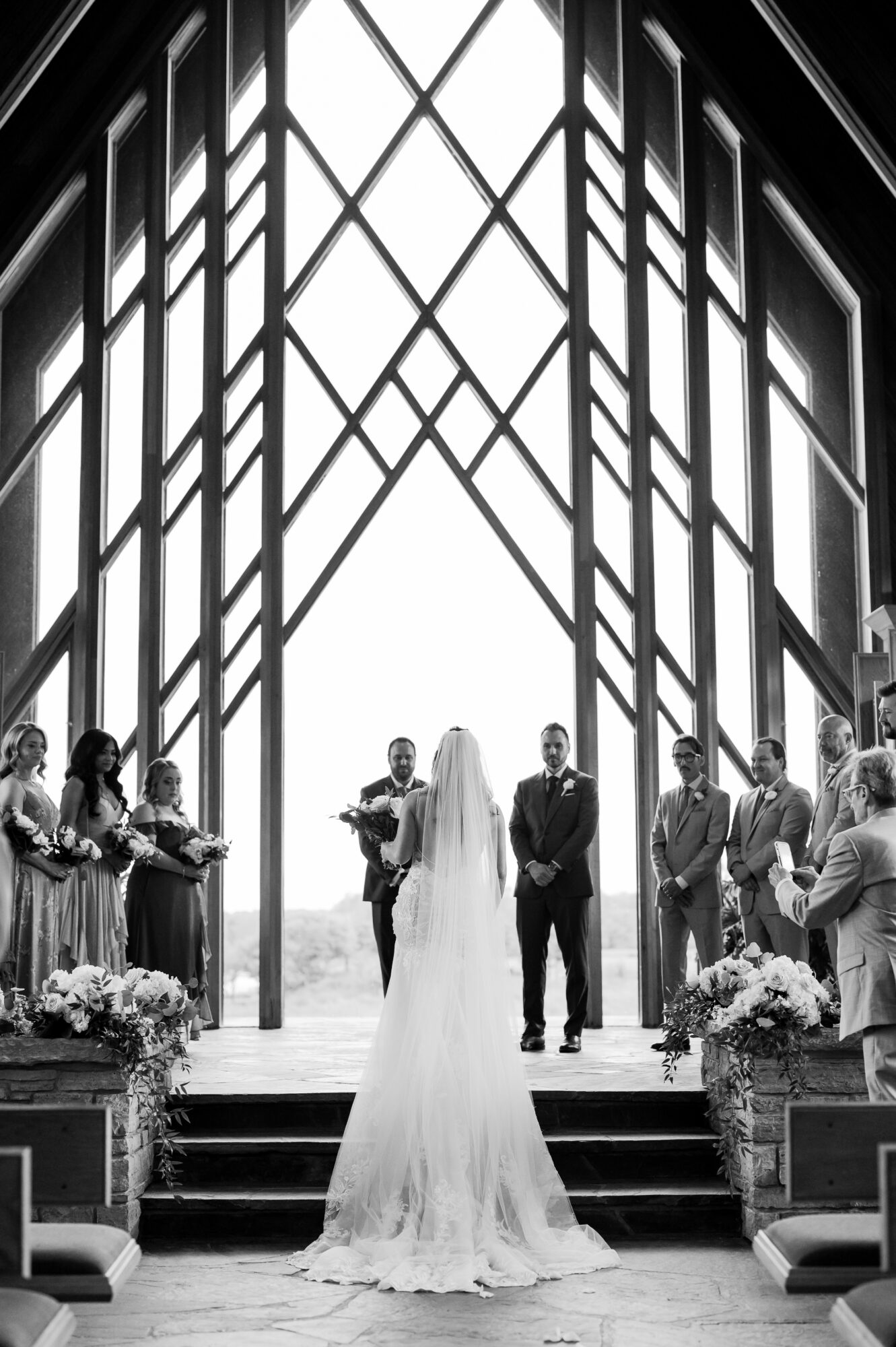 Bride and groom stand at altar with officiant and wedding party inside a modern glass chapel, floral arrangements on steps.