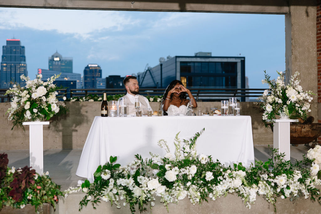 Two people sit at a decorated table with white flowers on a rooftop with city skyline background.