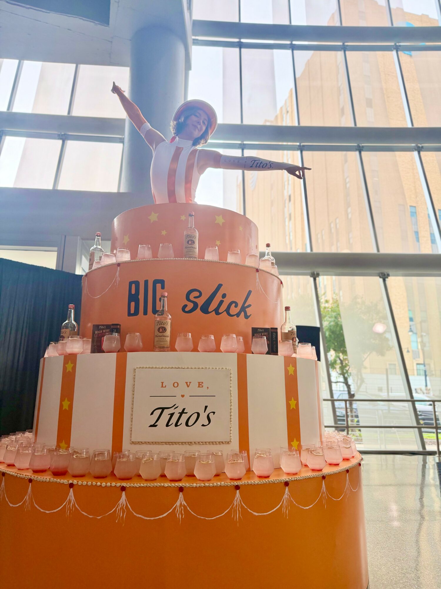 Cake with a figure of a person on top, decorated with bottles and a sign reading 'Tito's', inside a building with large windows.