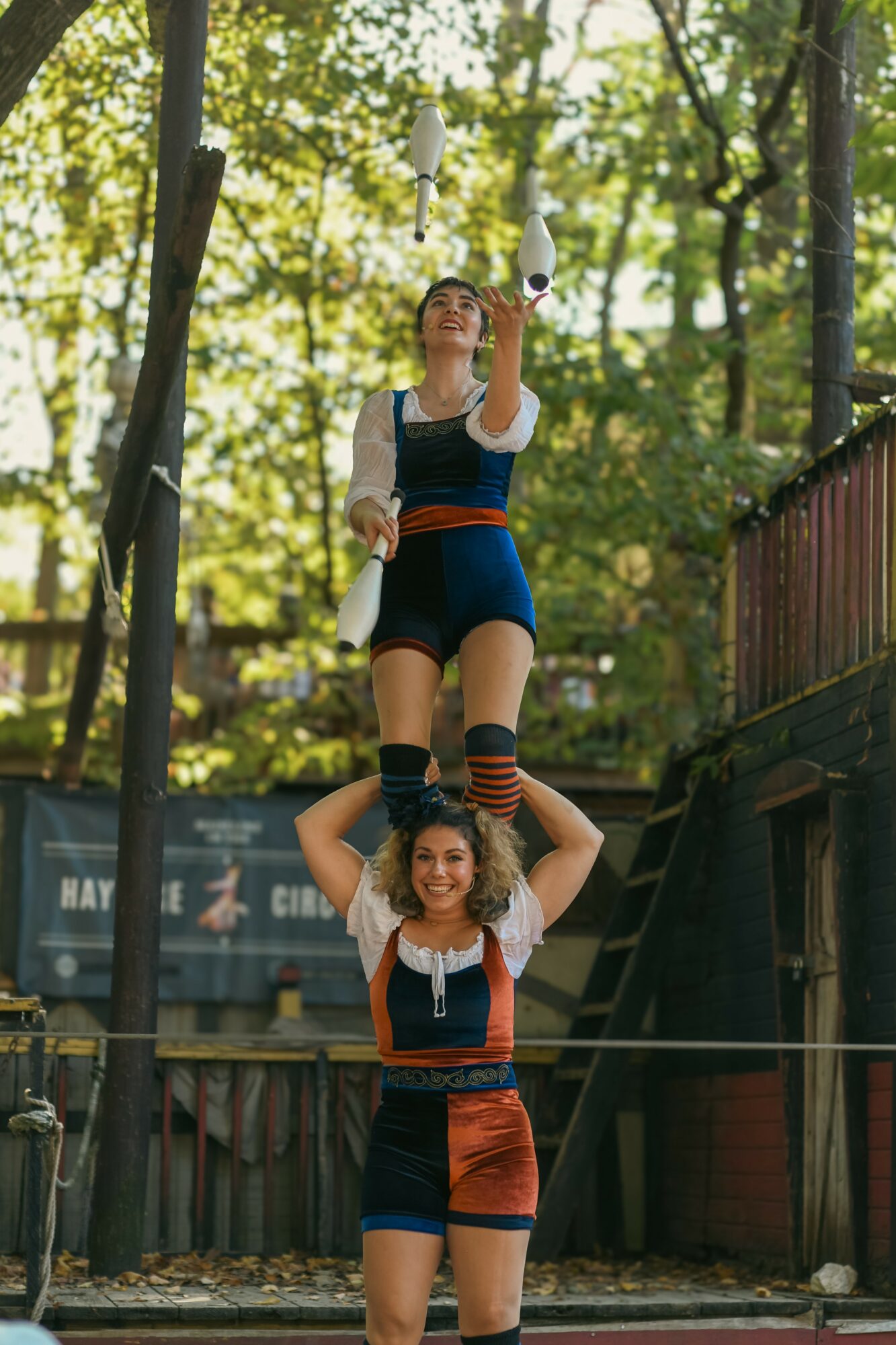 Two performers in traditional costumes balance on a tightrope outdoors among trees, with sunlight filtering through leaves.