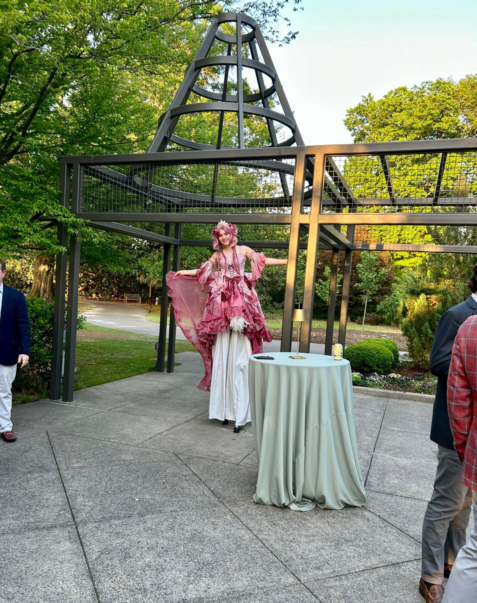 Person in pink dress standing on a white platform outdoors, with a round table nearby and trees in the background.