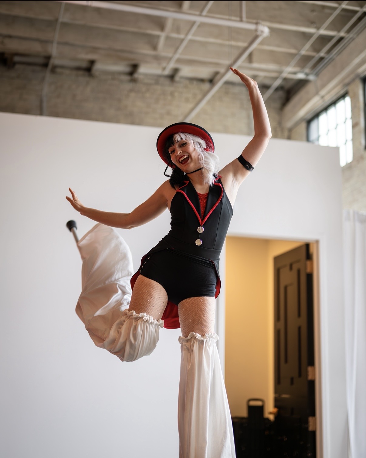 Person in a black outfit and red hat dancing with one leg raised, holding a stick, in a spacious room.