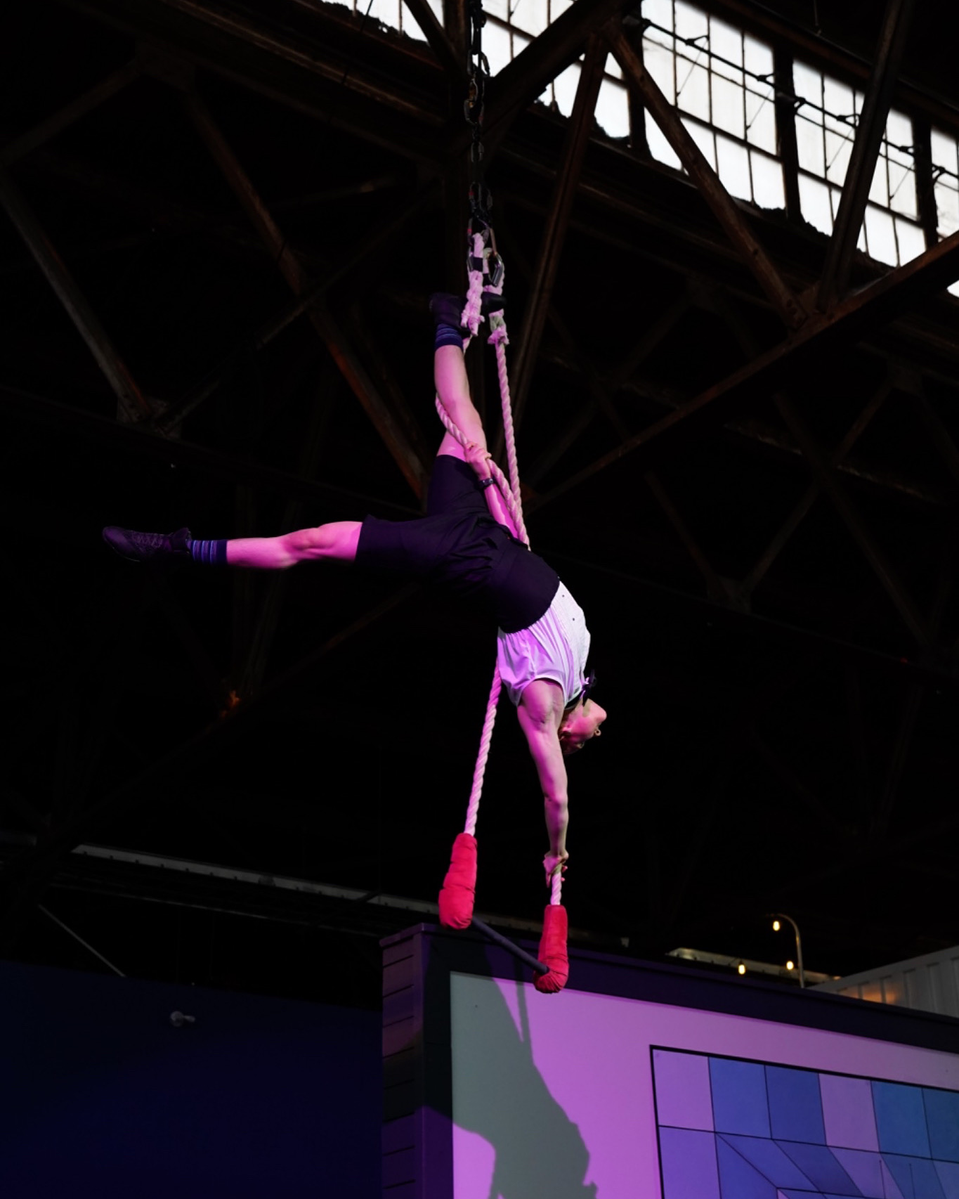 Person hanging upside down from a rope in an industrial setting, with a large window and metal structure above.