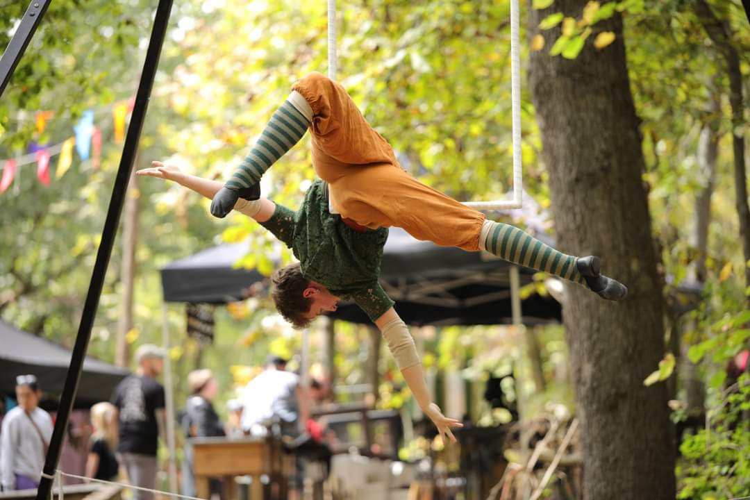 Person performing a handstand on a swing in a park with trees and people in the background.