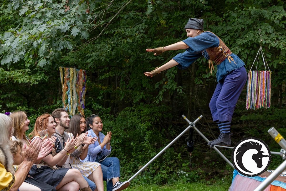 A performer balancing on a tall pole with arms outstretched, audience clapping outdoors with trees in background.