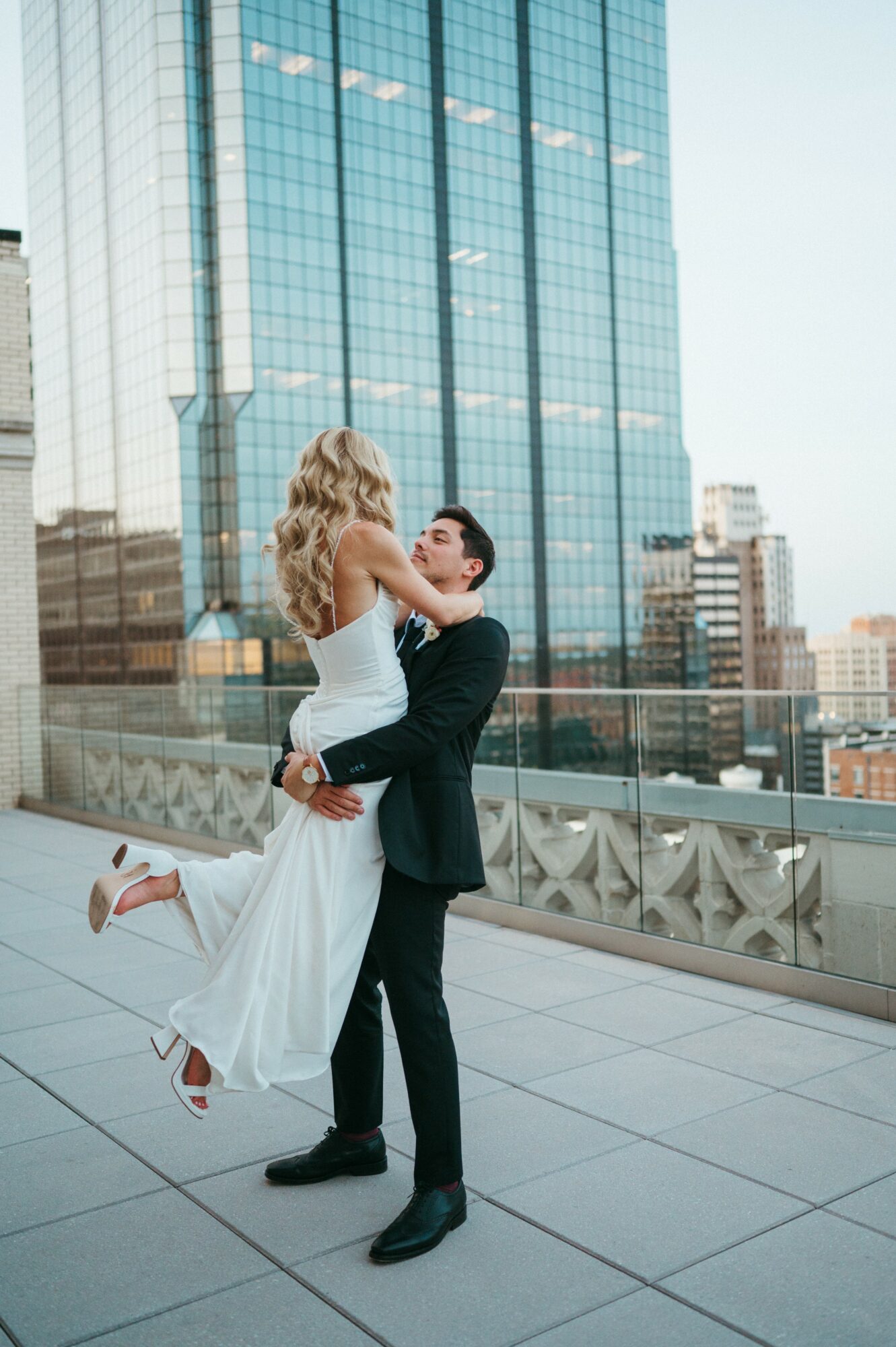 A couple dancing on a rooftop with city buildings in the background, the woman in a white dress and the man in a black suit.