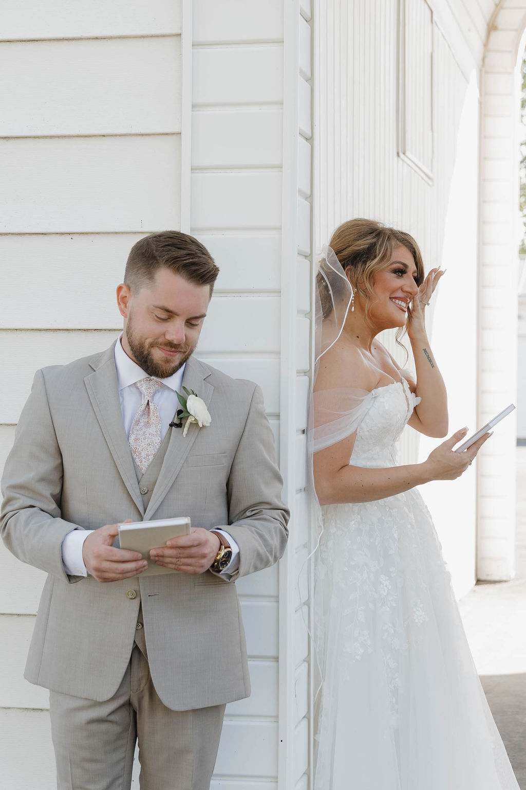 Bride in wedding dress laughing, groom in suit reading a card, outside white building, bright sunlight.