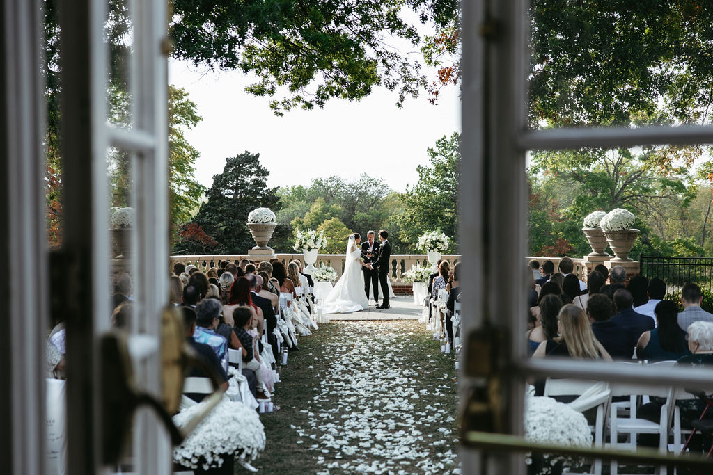 Wedding ceremony outdoors with guests seated, couple standing at altar, trees and decorative urns in background.