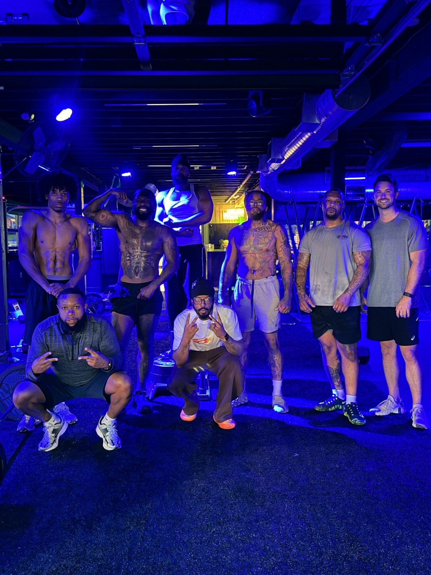 Group of people standing and sitting indoors with blue lighting, some wearing casual clothing, on a dark floor.