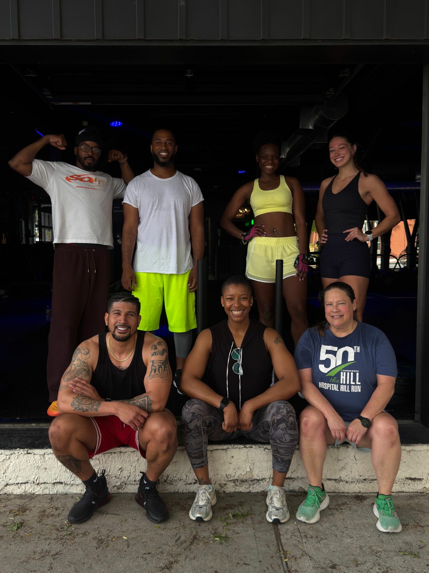 Group of people, some sitting and some standing, outdoors on a sidewalk, with a dark background and a person in a yellow vest.
