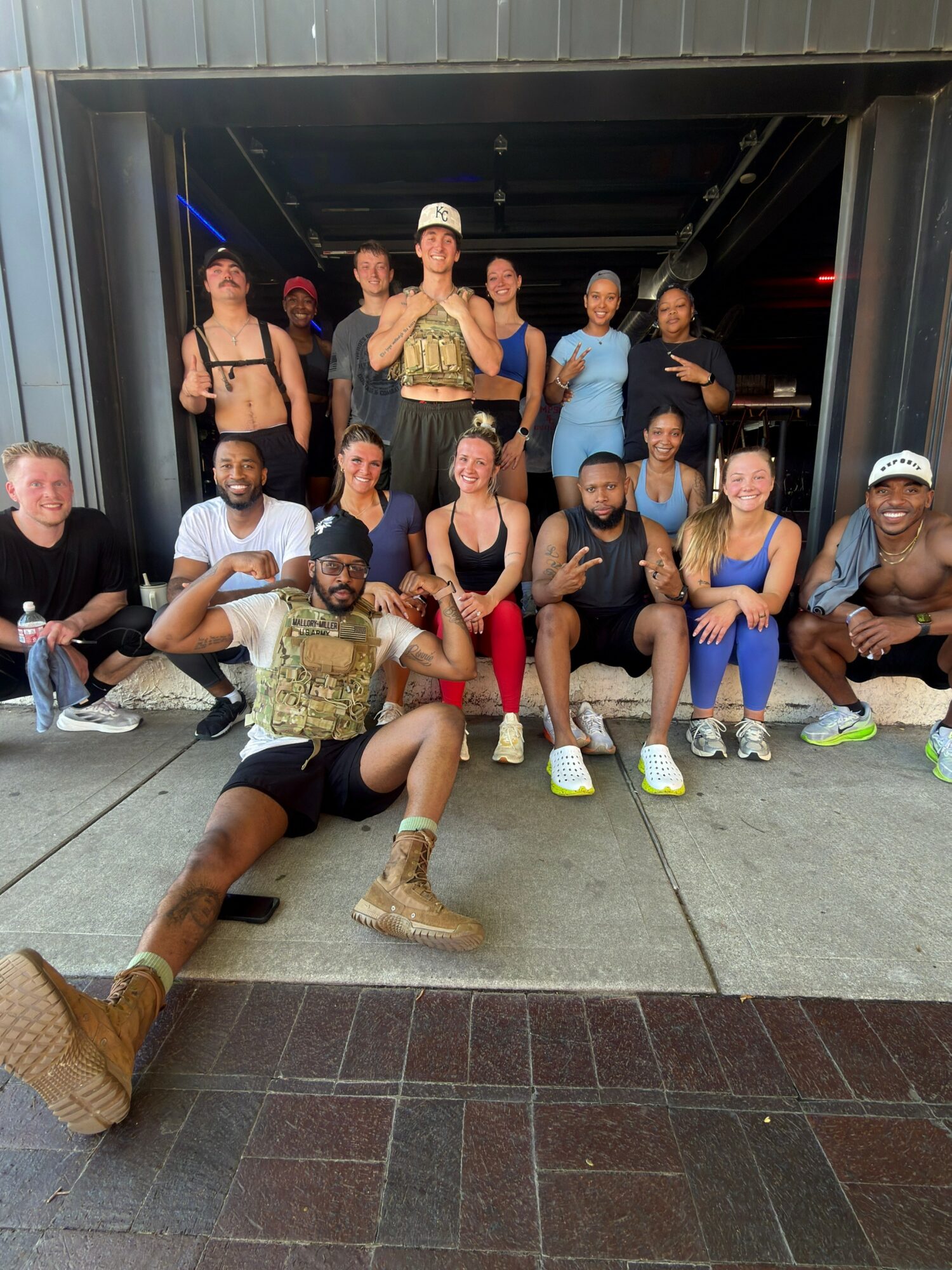 Group of people posing together outdoors, some sitting and some standing, in front of a dark structure, smiling and making gestures.