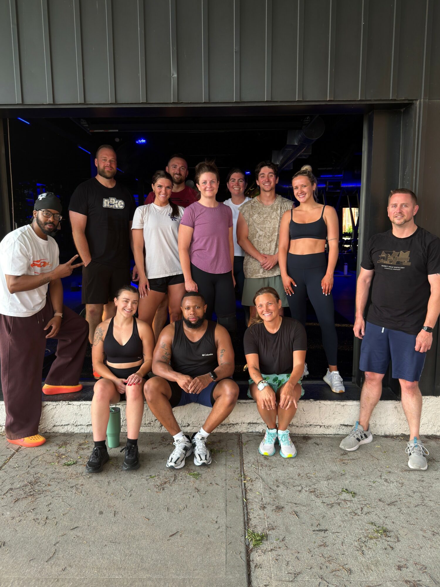 Group of ten people, some sitting and some standing, posing outside a building entrance. They are dressed casually, with some in athletic wear.